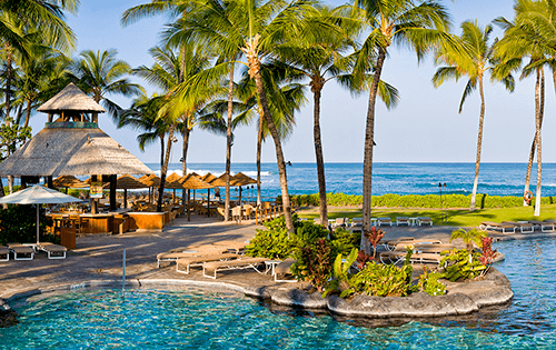 A blue pool on a Hawaiian resort with a beach and the ocean in the background.